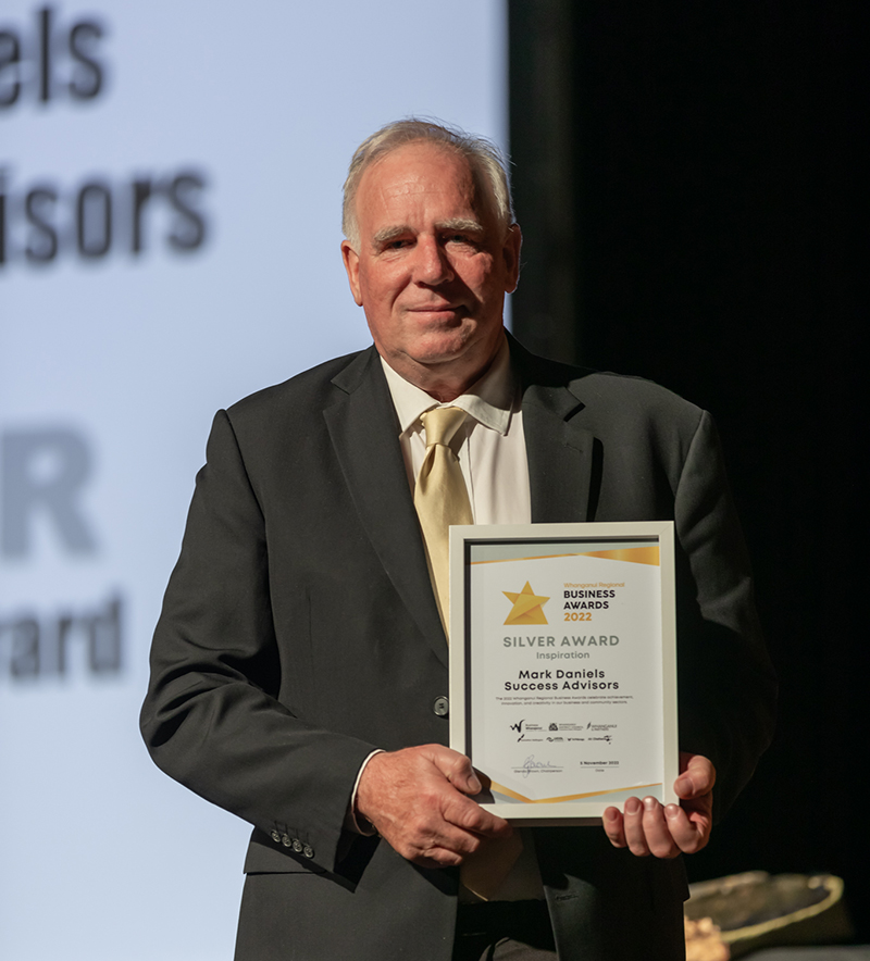 Mark Daniels holding an award. He is in a dark grey suit, gold tie and white shirt. The award is in a white picture frame