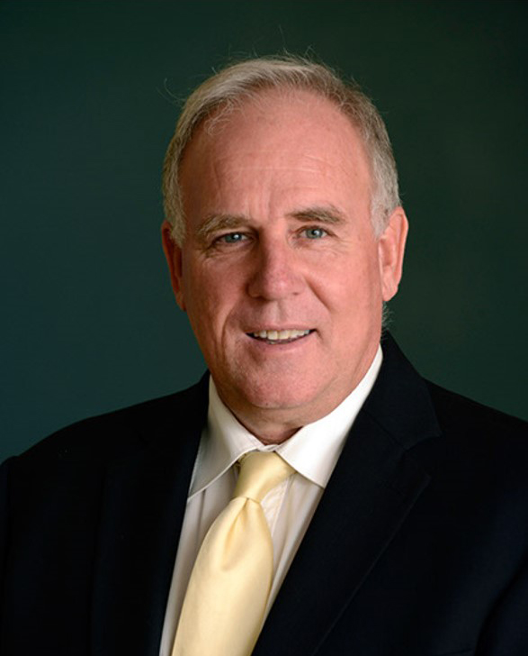 Portrait shot of Mark Daniels smiling at the camera. He is wearing a black suit, white shirt and gold tie.